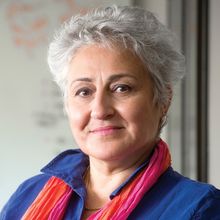 Haydeh Payami is wearing a purple dress and an orange and pink scarf and standing in front of a whiteboard.