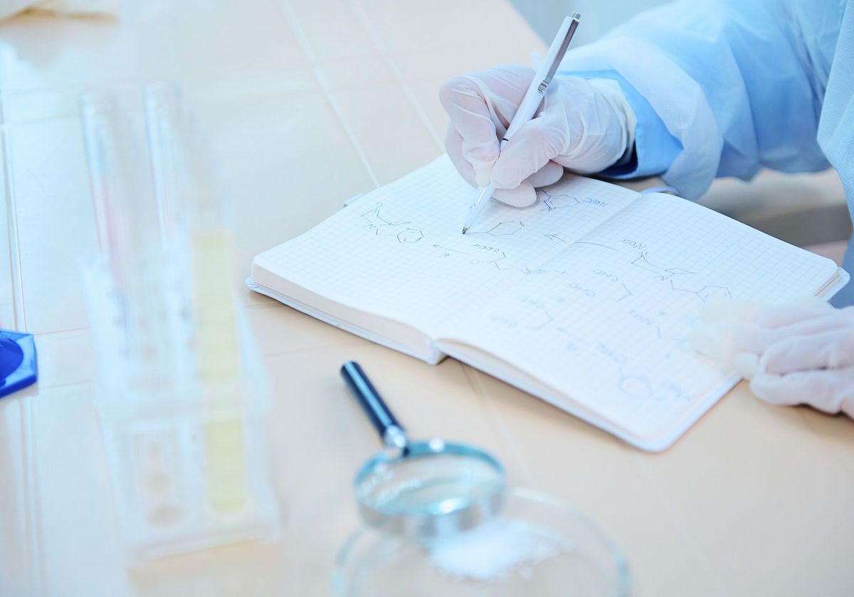 A scientist with gloved hands sitting at a table with a magnifying glass and lab glassware and writing in a notepad with a pen. 