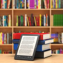 A tablet propped up by a stack of books in front of a bookshelf.