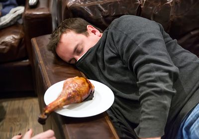 A photograph of a man sleeping on a couch next to a plate with a poultry leg on it.