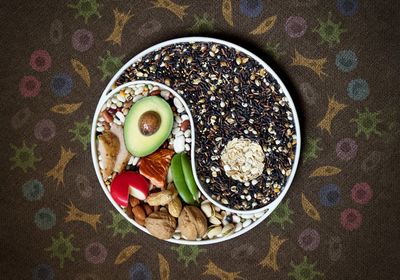 A bowl filled with various nuts, beans, seeds, avocado, oats, cheeses, and meat, is arranged by color to form a yin-yang symbol. The background is filled with cartoon images of cells.