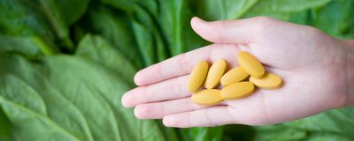 Image of a hand holding yellow pills in front of a leafy vegetable background.