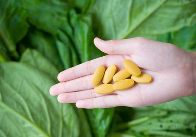 Image of a hand holding yellow pills in front of a leafy vegetable background.