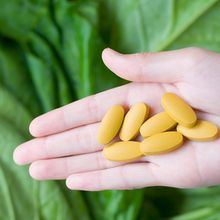 Image of a hand holding yellow pills in front of a leafy vegetable background.