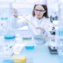 Scientist at lab bench holding a pipette with a microscope and glassware in the foreground.