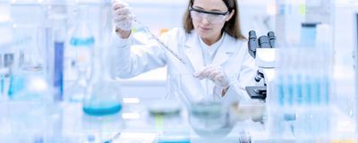 Scientist at lab bench holding a pipette with a microscope and glassware in the foreground.