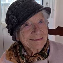 Older woman with hat and pink blouse smiling at camera.