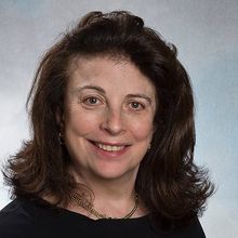 A portrait shot of Beryl Benacerraf, who wears a black shirt and gold necklace and smiles into the camera, on a bluish gray background.