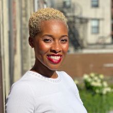 A Black woman stands in profile with her head turned towards the camera, smiling