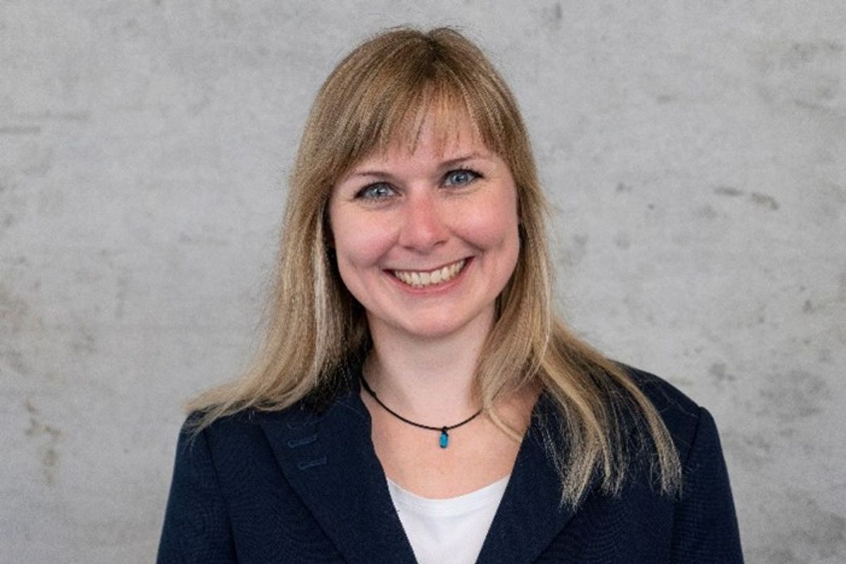 Scientist wearing a black blazer and white shirt smiling to the camera with a light grey wall in the background.