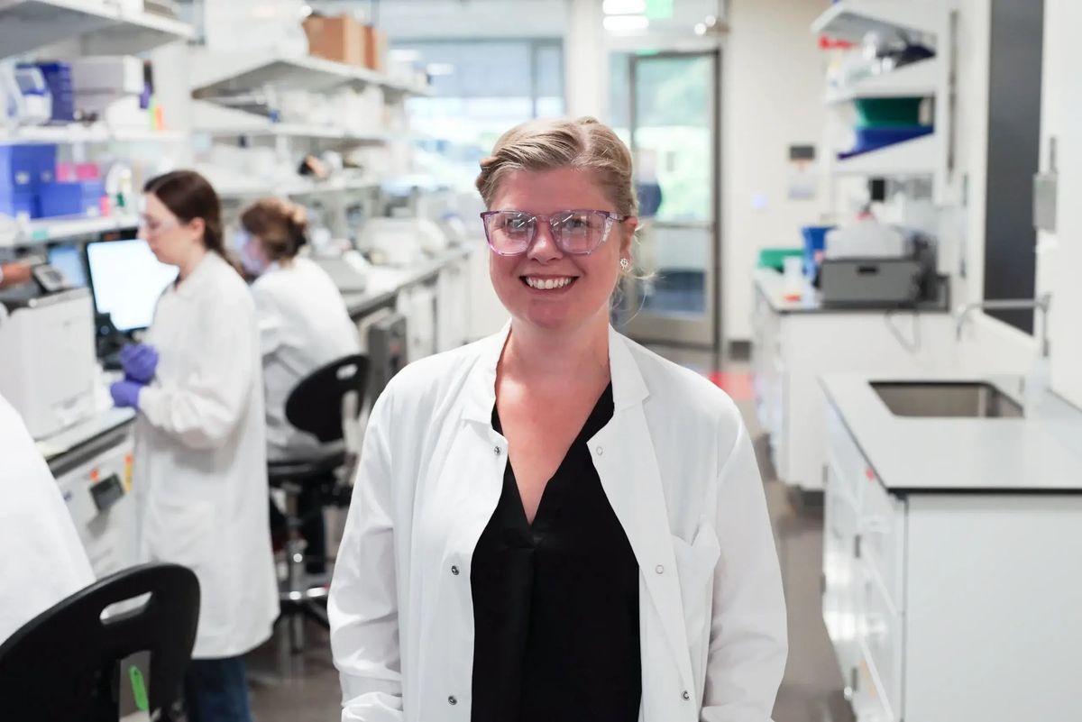 Image of immunologist Claire Gustafson. She wears glasses and smiles at the camera while wearing a black shirt under a white lab coat.