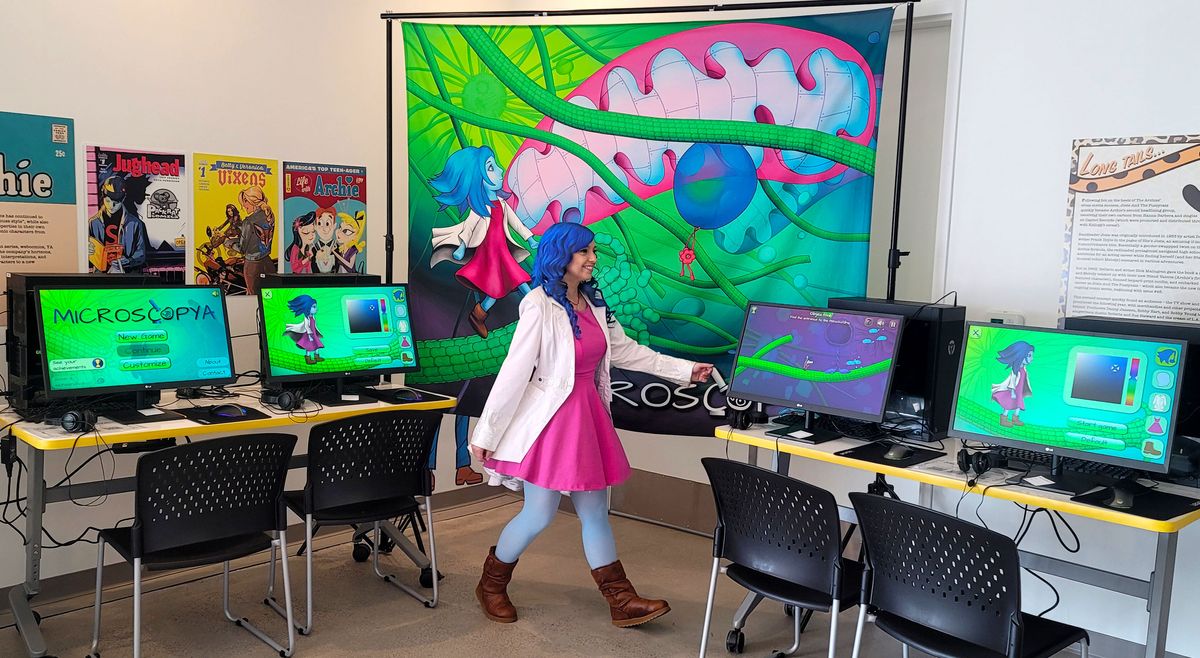 Beata Mierzwa, dressed in a pink dress, lab coat, and sporting blue hair, resembles the animated character from the game, Microscopya, she and her team designed. Computer stations beside her have the game pulled up at the 2022 San Diego Comic-Con.