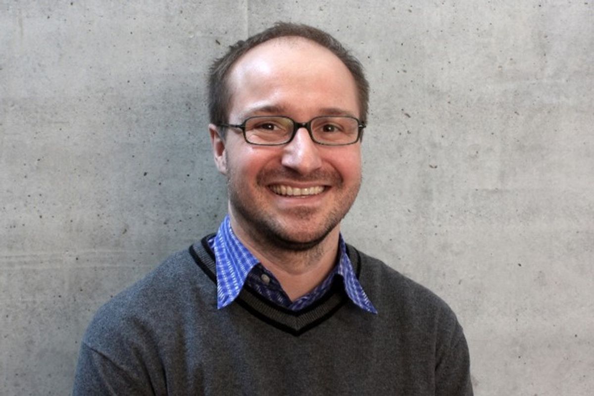 Scientist wearing a dark grey sweater, blue checkered shirt, and glasses smiling to the camera with a light grey wall in the background.