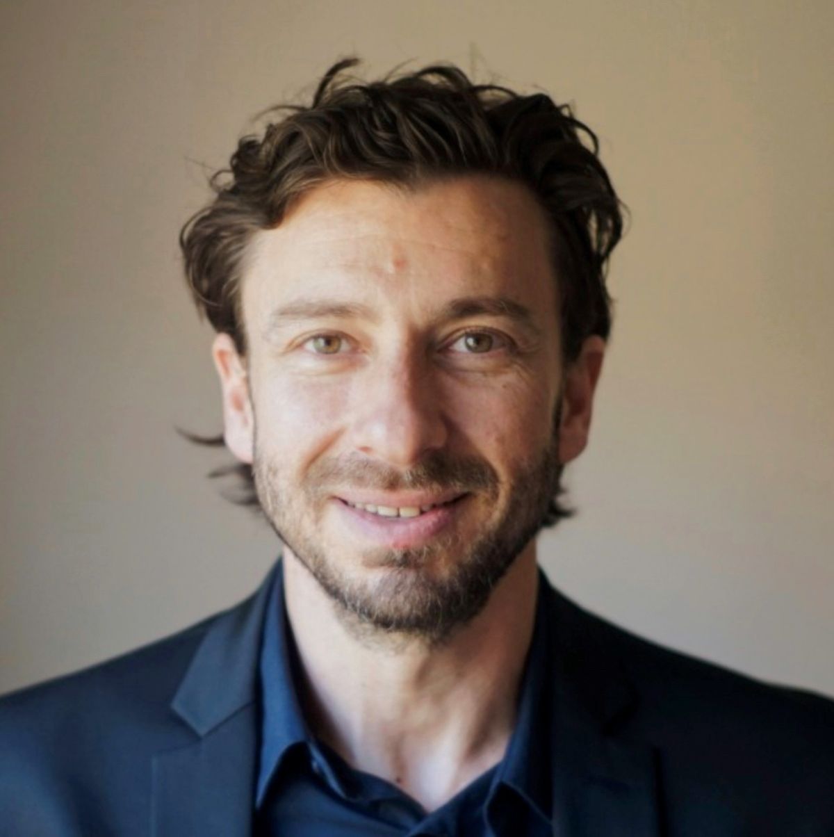 A photograph of David Furman, an immunologist and data scientist at the Buck Institute and Stanford University. Furman has dark brown hair, wears a dark blue dress shirt and jacket, and is looking at the camera.