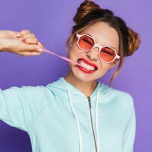 Woman with buns and blue sweater chewing gum, smiling, and stretching it out of her mouth.