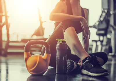 A person sitting in a gym with a protein shake bottle and kettlebell placed beside them.