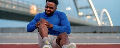 A man holds his knee and grimaces in pain while sitting in front of a bridge.