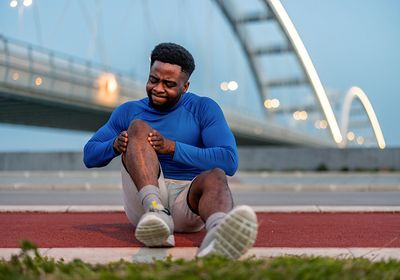 A man holds his knee and grimaces in pain while sitting in front of a bridge.