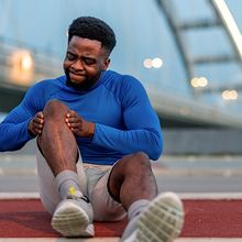 A man holds his knee and grimaces in pain while sitting in front of a bridge.