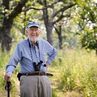 Dr. Brock standing in nature, holding a walking stick and wearing binoculars around his neck.